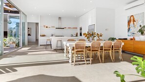 A modern kitchen with white cabinetry, a dining area with a white table and wicker chairs, and a large window providing natural light. - POOLSIDE PARADISE - MOUNT ELIZA (Mount Eliza)