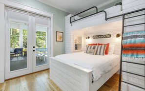 Une chambre avec un lit superposé, une porte vitrée donnant sur un patio extérieur et une enseigne fixée au mur sur laquelle on peut lire « MAISON DE PLAGE ».