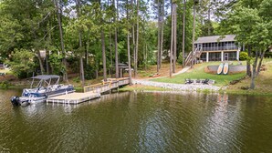 A lakeside property with a dock, boats, a covered boat house, a house with a deck, and a grassy area with outdoor seating. - Home w/Dock, SUPs, Kayaks, Arcades, Ping-Pong (Leesville)