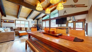 A spacious living room with a wooden dining table, benches, and a sofa. There is a fireplace, a television, and large windows allowing natural light. - Chateau Sans Nom #19 4 Bedroom Condo by RedAwning (Mammoth Lakes)