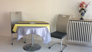 A dining area with a table covered by a yellow and white tablecloth, a chair, and a vase with flowers on a radiator. - Charmant Studio 42 M2 10 min de la Plage (Le Cannet)