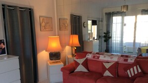 A living room with a red sofa, two bedside lamps, a small table, and a window with curtains. - Charmant Studio 42 M2 10 min de la Plage (Le Cannet)