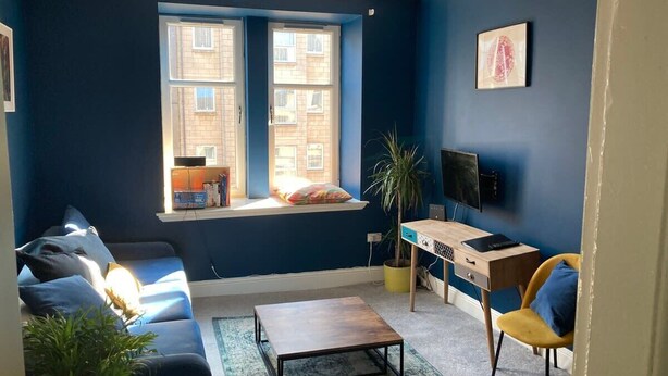 Ein Wohnzimmer mit einer Couch, einem Couchtisch, einem Schreibtisch mit Stuhl, einem Fernseher und einem Fenster mit Blick auf Gebäude.