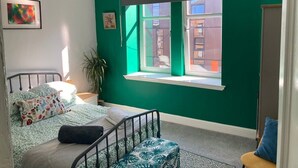 Ein Schlafzimmer mit grüner Wand, einem Bett mit gemusterten Kissen, einem Fenster mit Blick auf Gebäude und einer Pflanze.