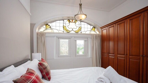 A hotel room with a large bed, two windows with curtains, a wooden wardrobe, and a chandelier.