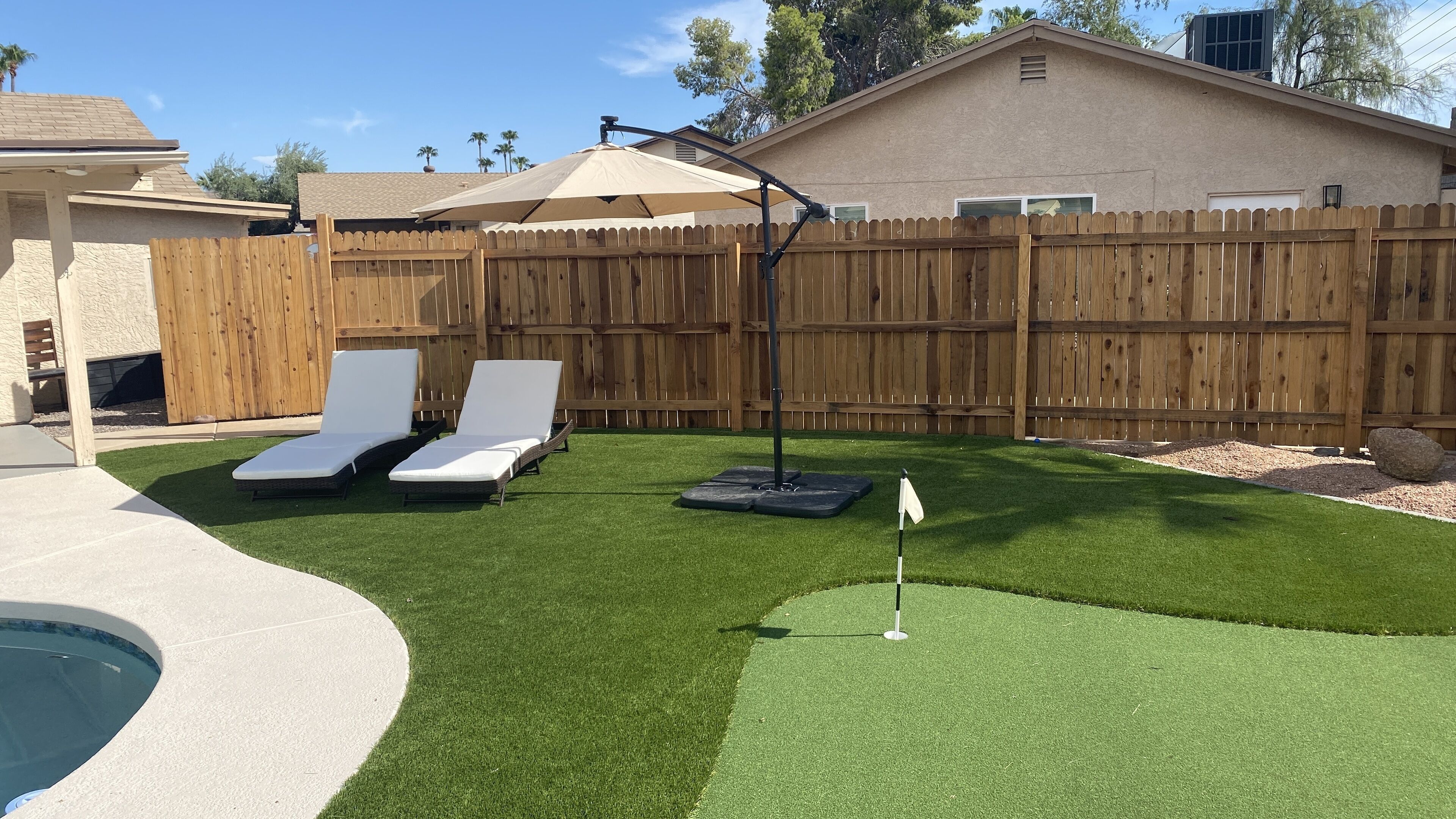 A backyard with artificial grass, two lounge chairs, a pool, and a wooden fence.