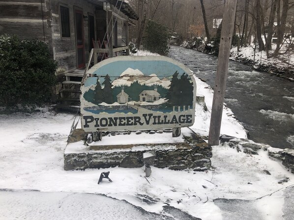 A Pioneer Village sign with a rustic wooden house and a river in the background.