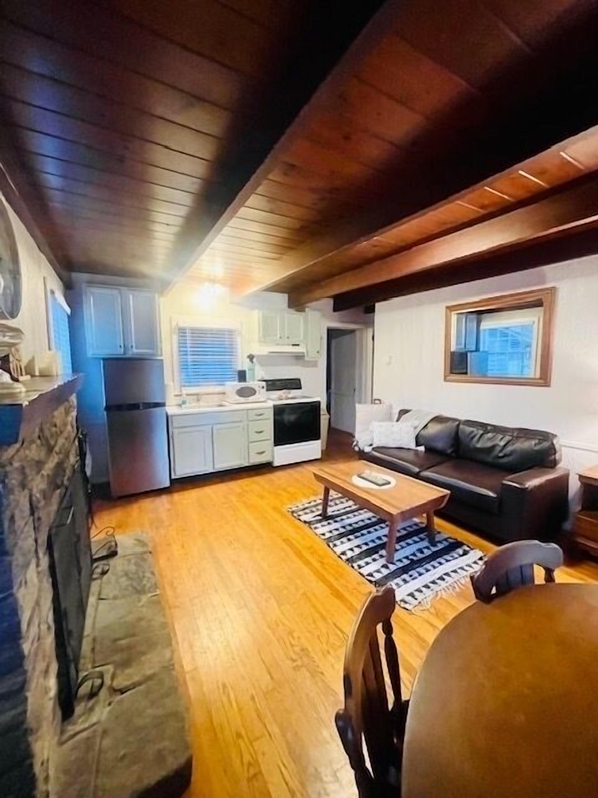 A cozy living room with a wooden floor, a black leather sofa, a coffee table, and a dining table with chairs. There is a kitchen area with white cabinets and a window.