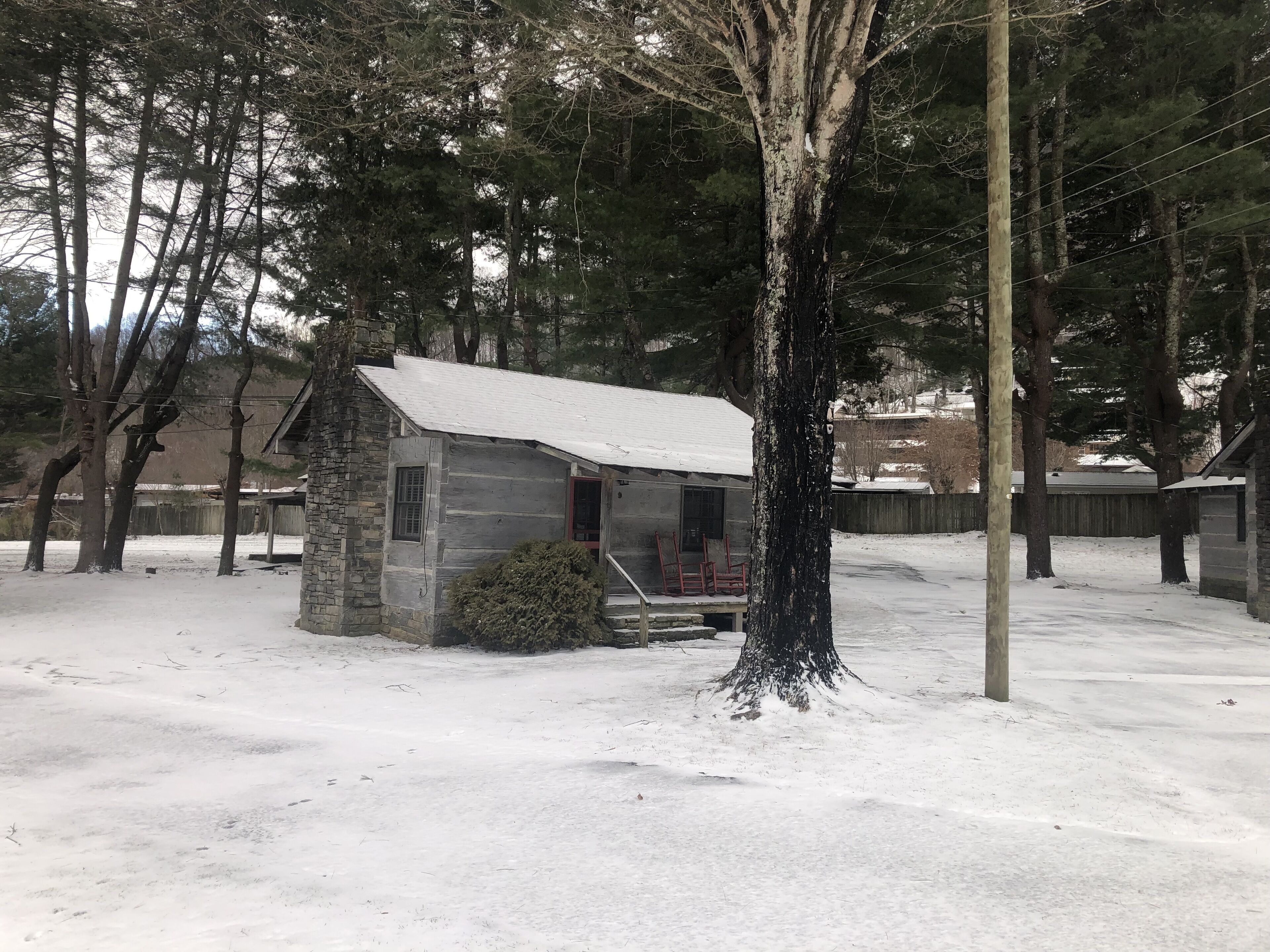 A small stone cabin with a snow-covered roof, surrounded by trees and a wooden fence.