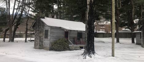 A small stone cabin with a snow-covered roof, surrounded by trees and a wooden fence.