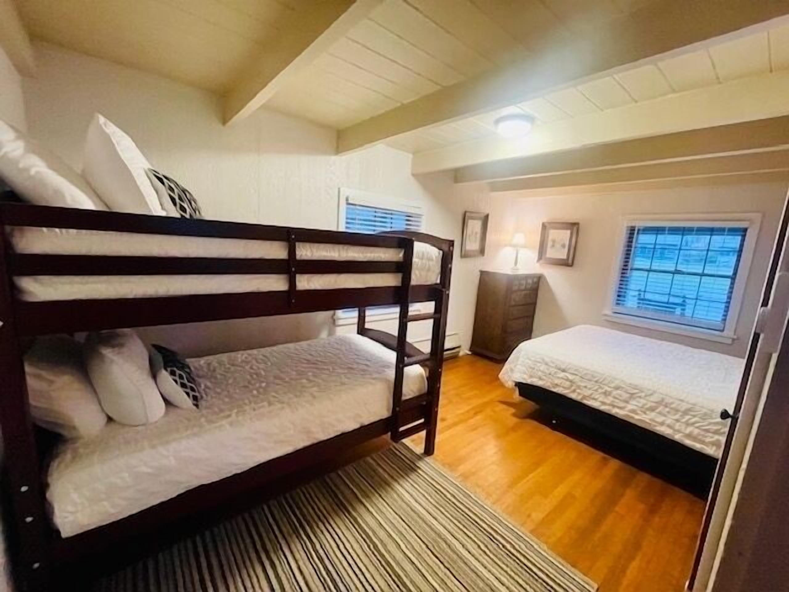 Een slaapkamer met een stapelbed, een enkel bed, houten vloeren, een raam met zonwering en een klein kastje.