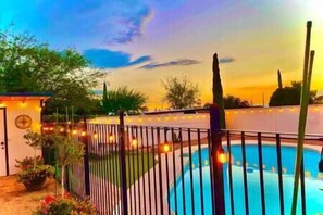 Una piscina con cerramiento metálico, rodeada de plantas en macetas y un pequeño edificio con luces colgantes.