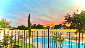 Ein Schwimmbecken, umgeben von einem Zaun mit Lichterketten, einem kleinen Gartenbereich und einem Sonnenuntergang im Hintergrund.