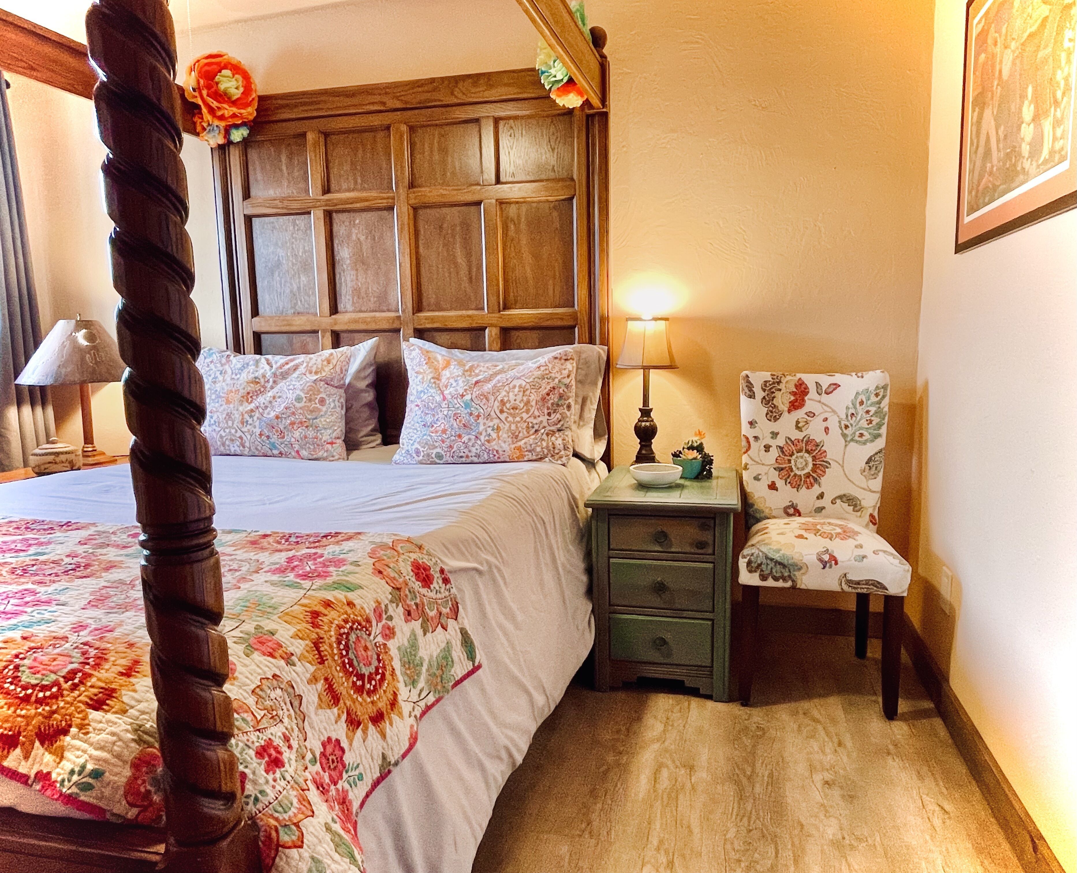 Une chambre à coucher avec un lit à baldaquin, une literie à motifs floraux, une table de chevet avec une lampe, un fauteuil à motifs floraux et une tête de lit en bois.
