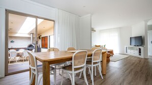 Una sala de estar moderna con una mesa de comedor de madera, sillas de mimbre blanco, un televisor de pantalla plana y un gran ventanal corredizo que da a un balcón.