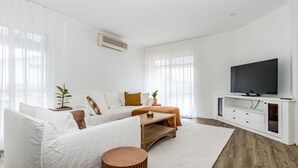 Un salón moderno con un sofá blanco, una mesa de centro, un televisor sobre un mueble blanco y una planta en maceta.