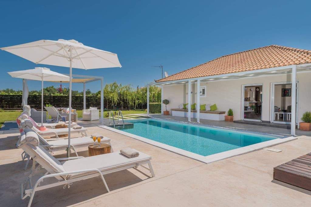 Une piscine avec des chaises longues et des parasols, un espace de détente couvert et une maison avec un toit en tuiles.
