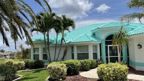A building with a turquoise roof, surrounded by well-maintained landscaping and palm trees. - Family Canal House/ Heated Pool, Kayaks, BBQ, Lift (Cape Coral)