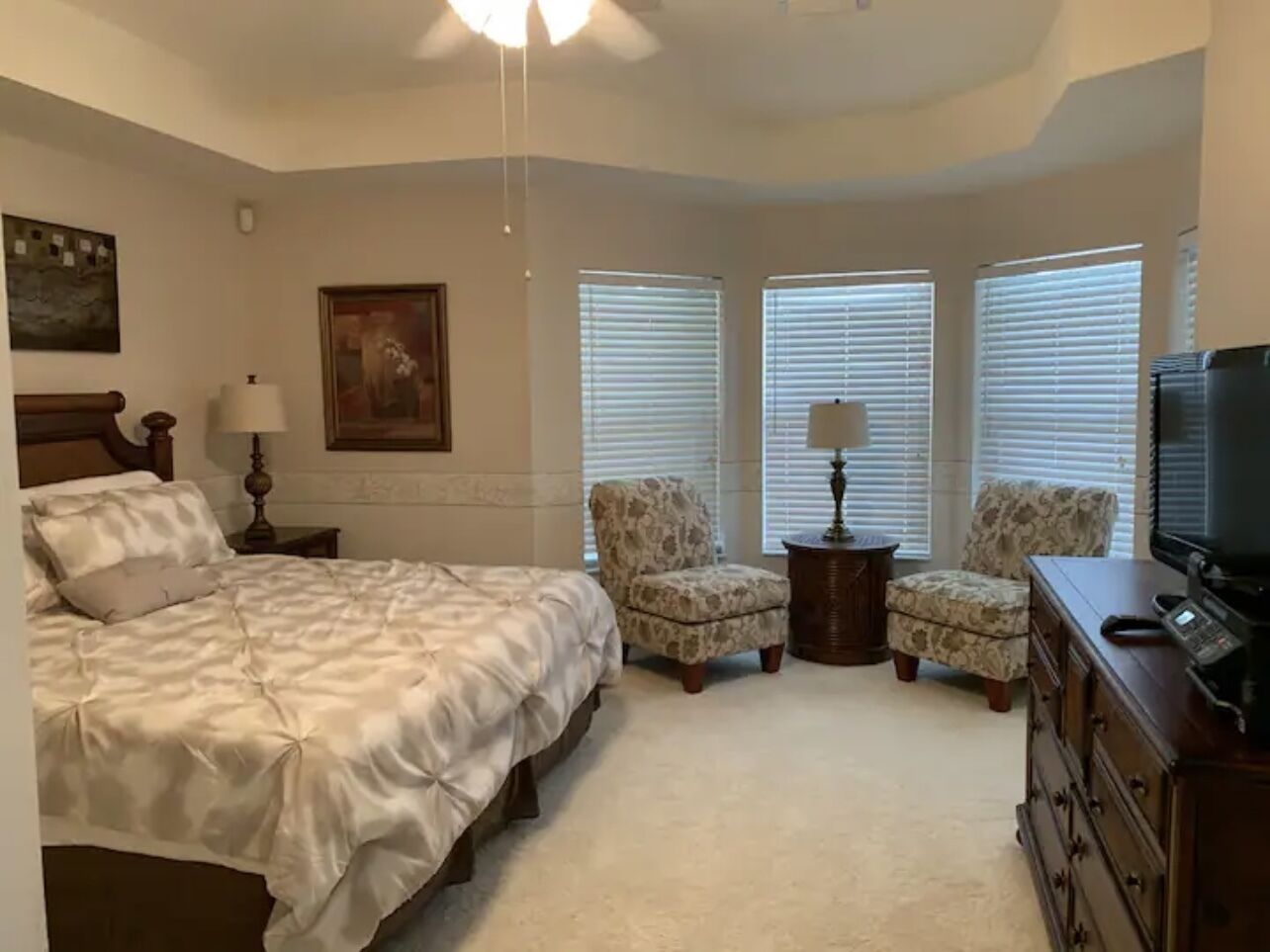 A bedroom with a large bed, two armchairs, a dresser, a television, and a ceiling fan.