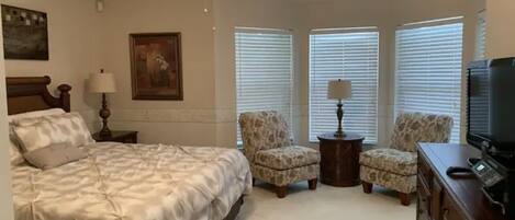 A bedroom with a large bed, two armchairs, a dresser, a television, and a ceiling fan.