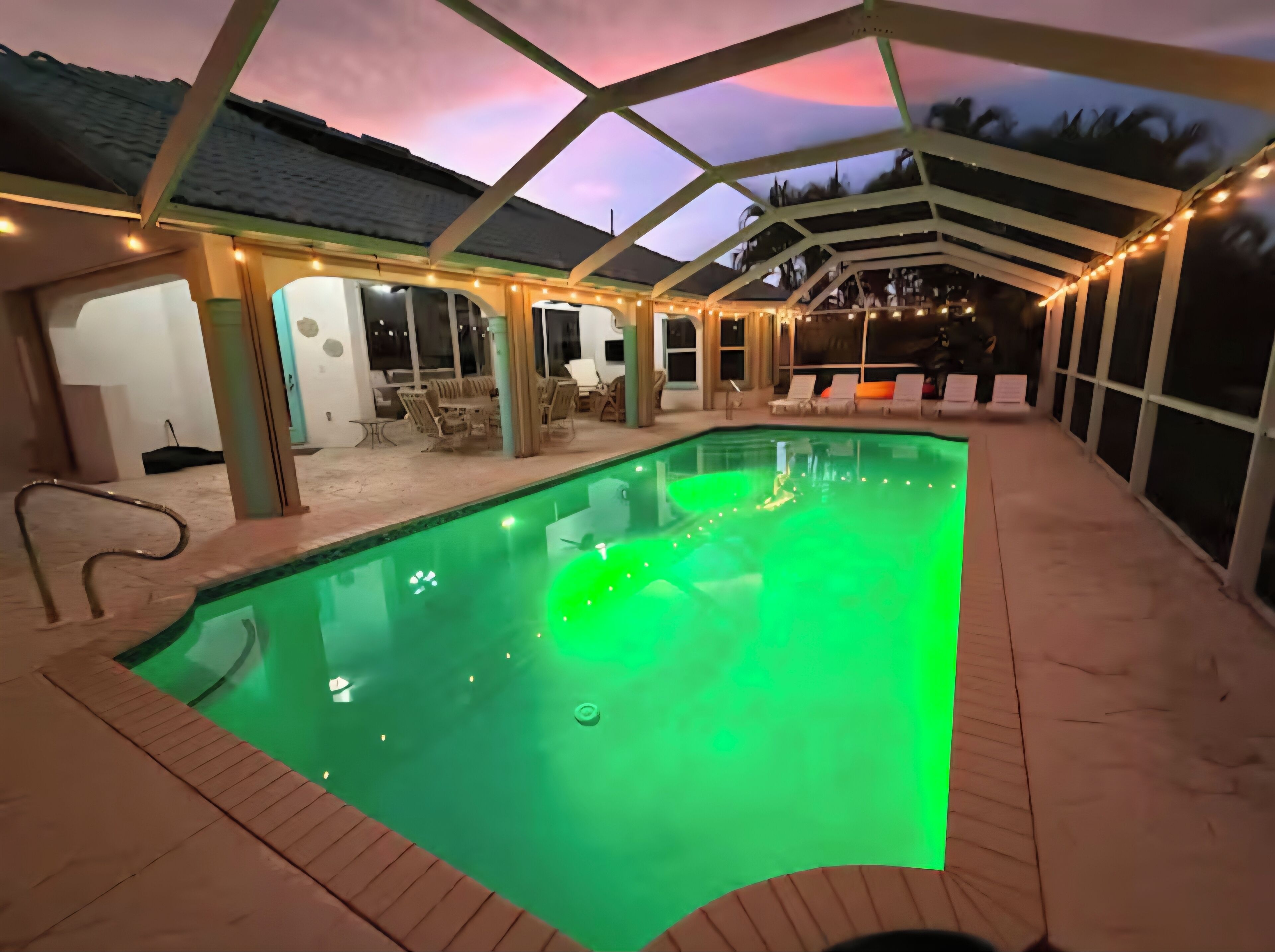 Ein Schwimmbecken mit grüner Beleuchtung, umgeben von einer überdachten Terrasse mit Stühlen und Tischen.