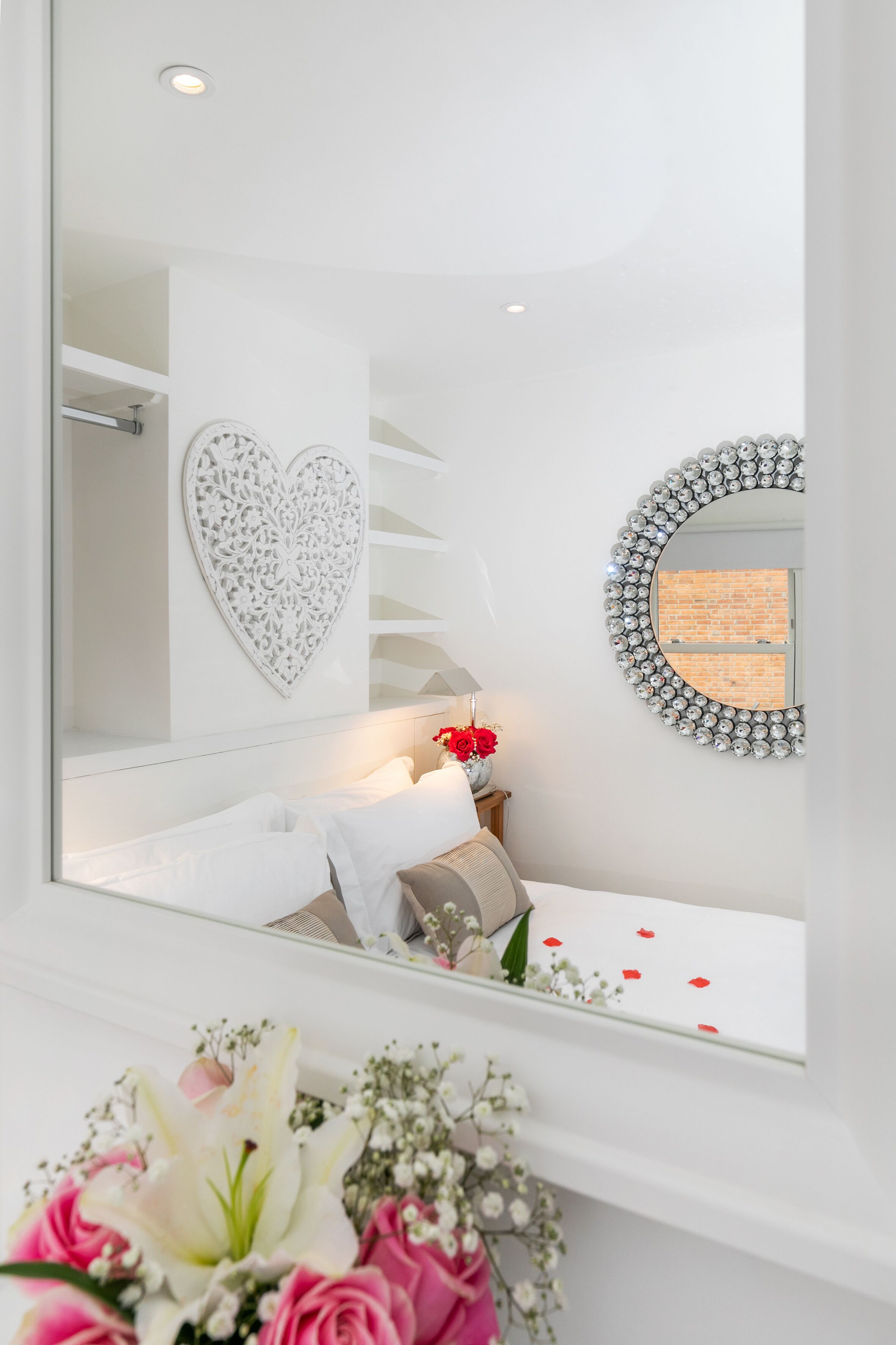A bedroom with a white bed, a heart-shaped wall decoration, a round mirror, and a window with a brick wall view.