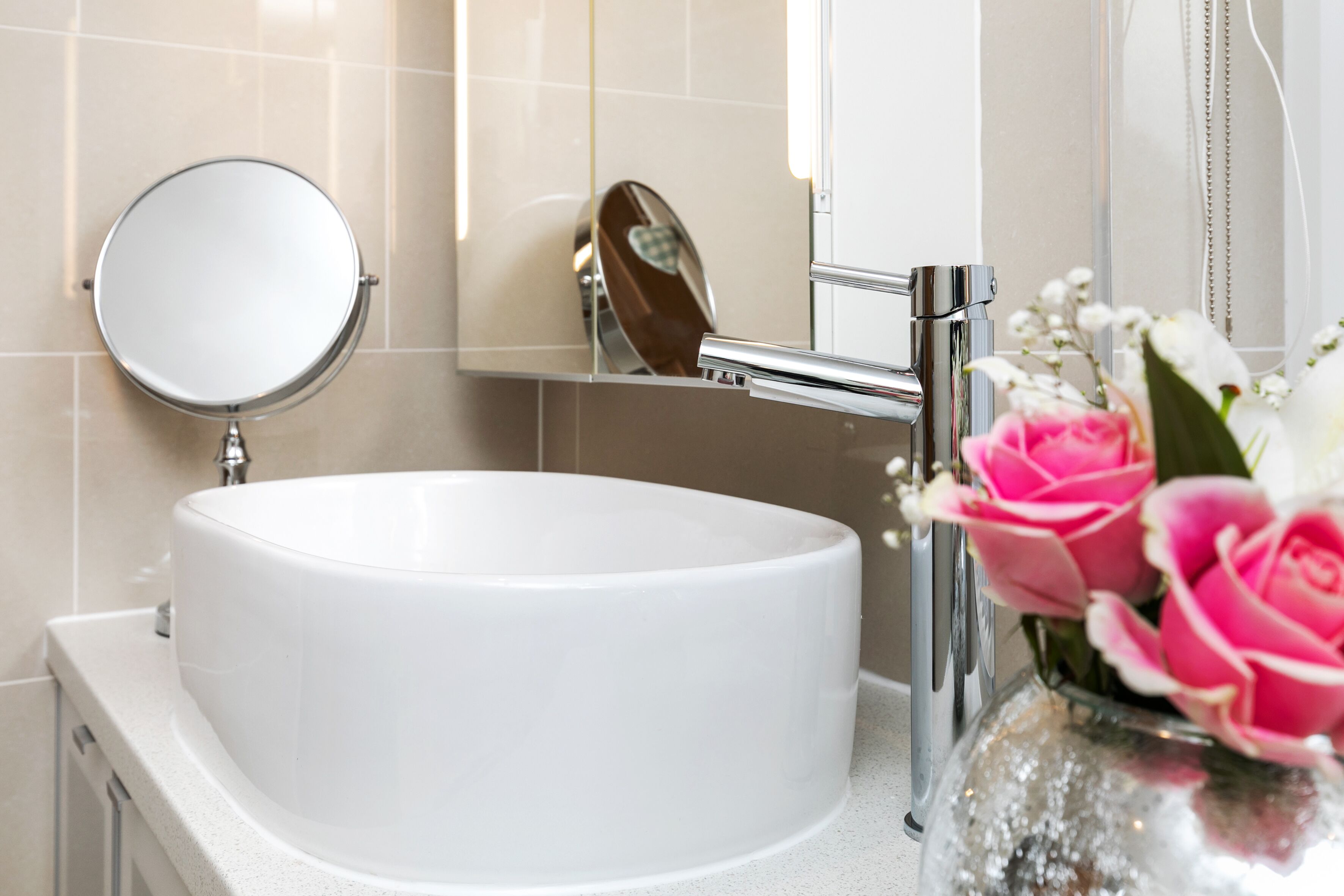 A bathroom with a round white sink, a mirror, and a vase with flowers.