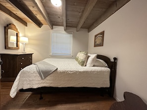 A bedroom with a wooden ceiling, a bed with a white quilt, a nightstand with a lamp, and a window with blinds.