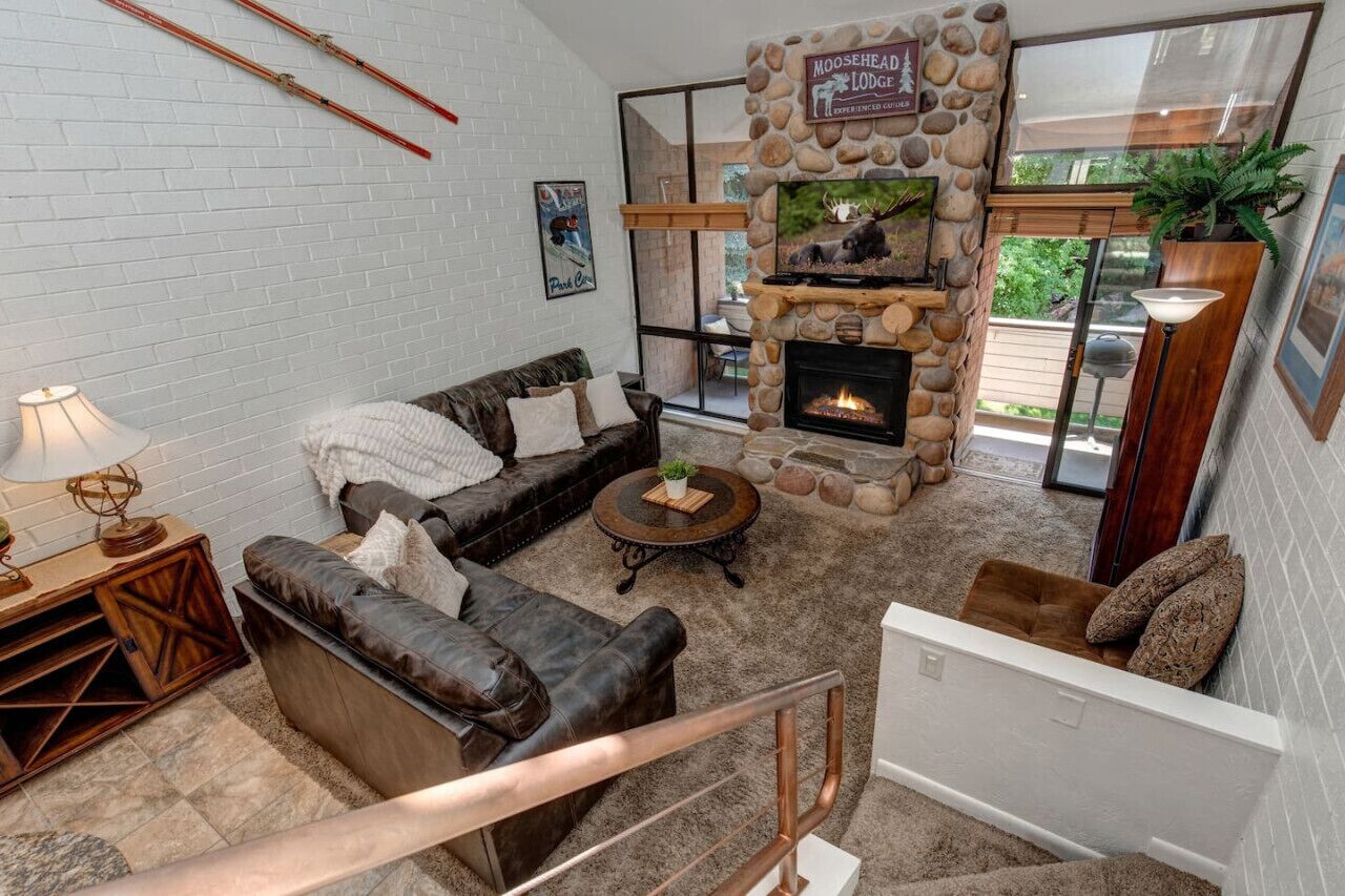 A cozy living room with a stone fireplace, leather sofas, and a large window offering a view of the outdoors.