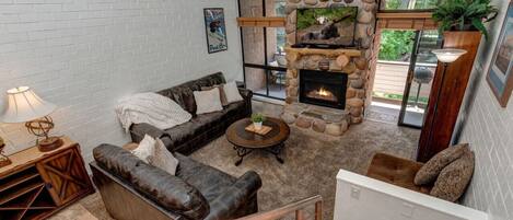 A cozy living room with a stone fireplace, leather sofas, and a large window offering a view of the outdoors.