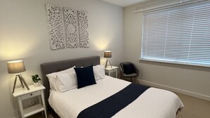 A neatly arranged bedroom with a large bed, bedside lamps, decorative wall art, and a window with blinds.