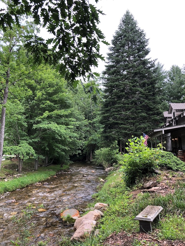 A serene riverside setting with a wooden cabin, lush greenery, and a small bench.