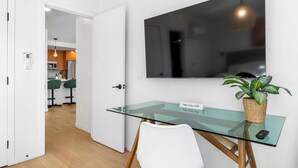 A modern room with a glass desk, a mounted TV, a potted plant, and a door leading to a kitchen area.