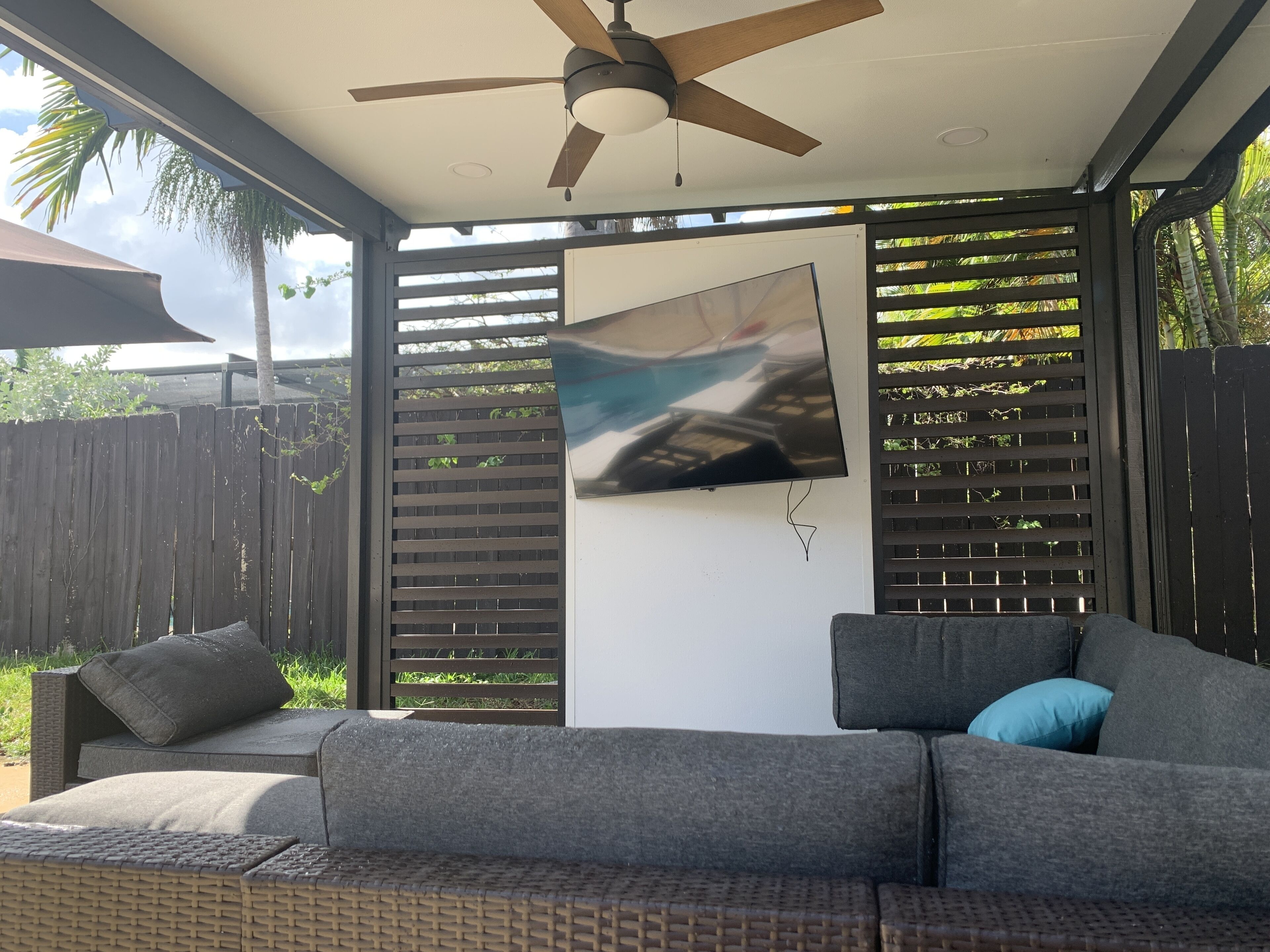 A covered outdoor seating area with a ceiling fan, a flat-screen TV mounted on the wall, and a wooden fence in the background.