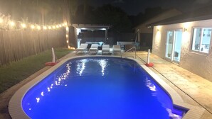 En swimmingpool med buet kant, omgivet af en træterrasse og en overdækket patio med udendørs møbler.