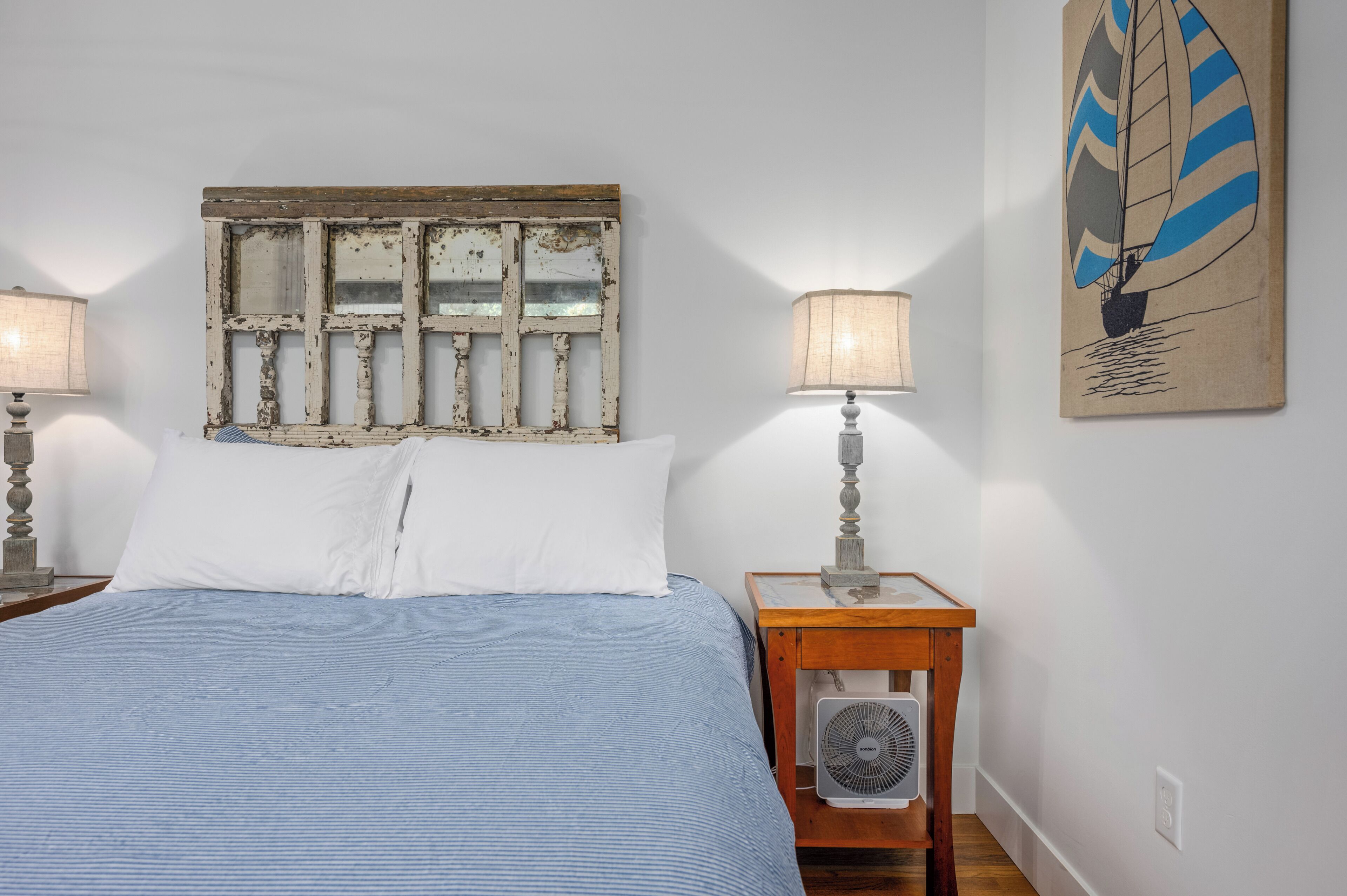 A bedroom with a bed, a wooden headboard, a wall-mounted fan, and a painting of a sailboat.