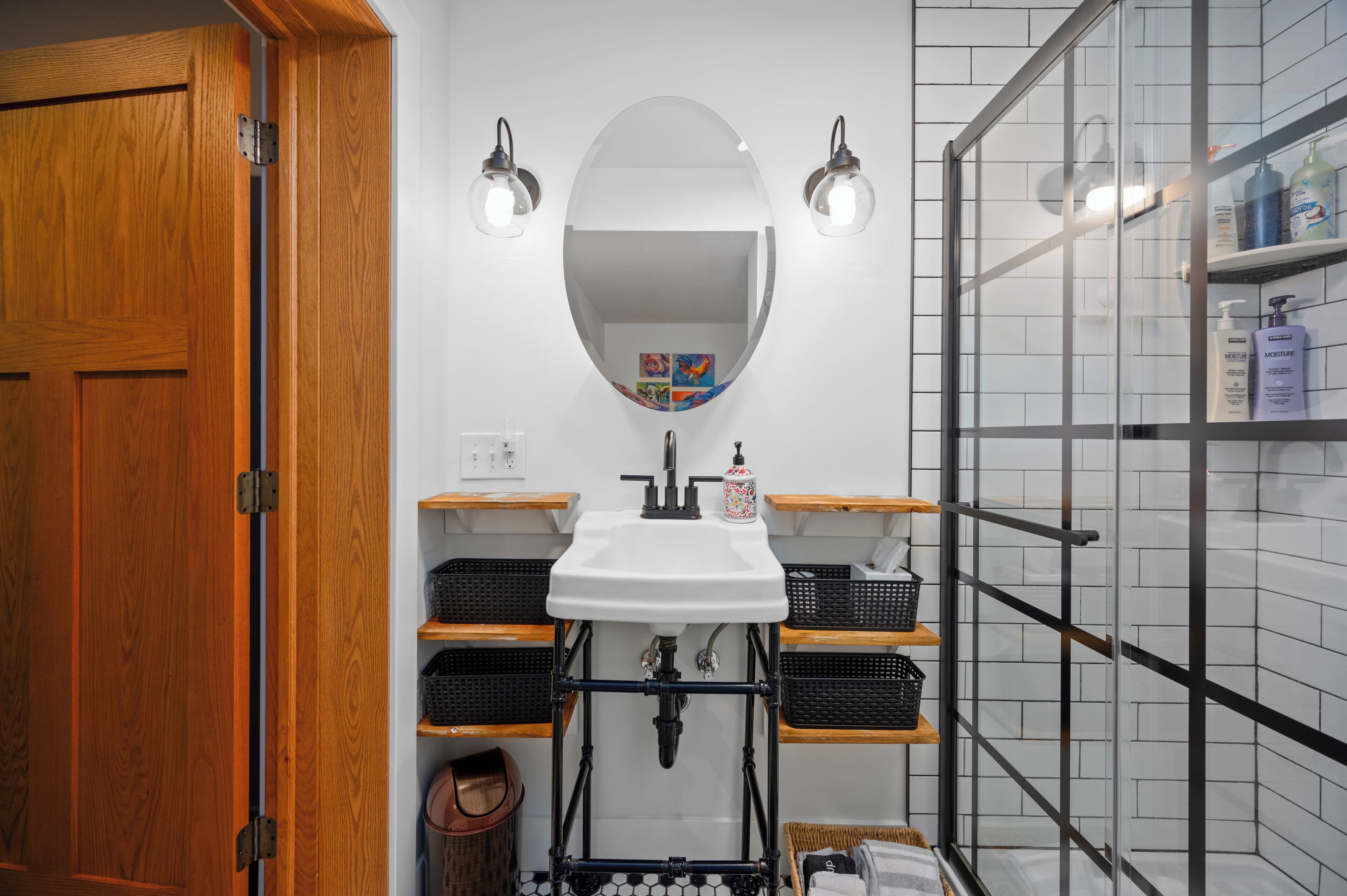 Et badeværelse med en trædør, en hvid vask med spejl, to vægmonterede lamper og hylder med toiletartikler.