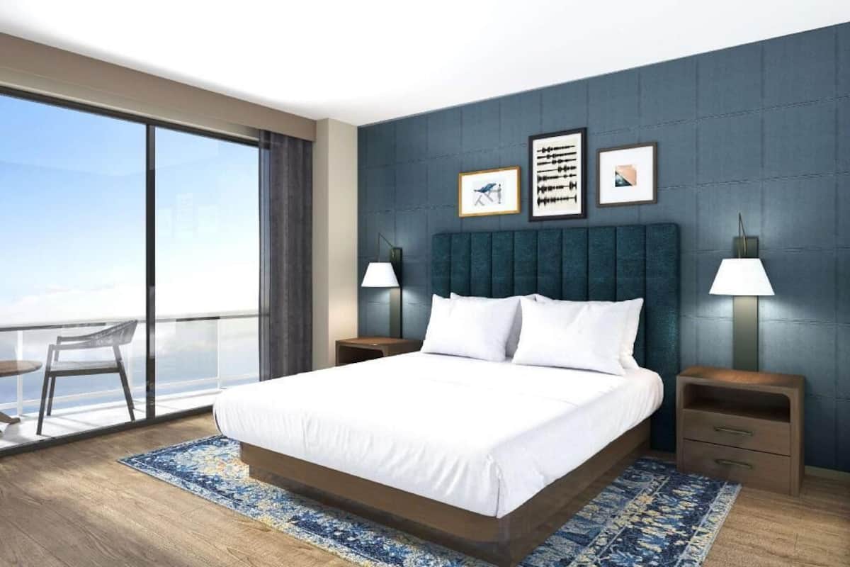 A modern hotel room with a large bed, two bedside tables with lamps, a balcony with a chair, and a city view.