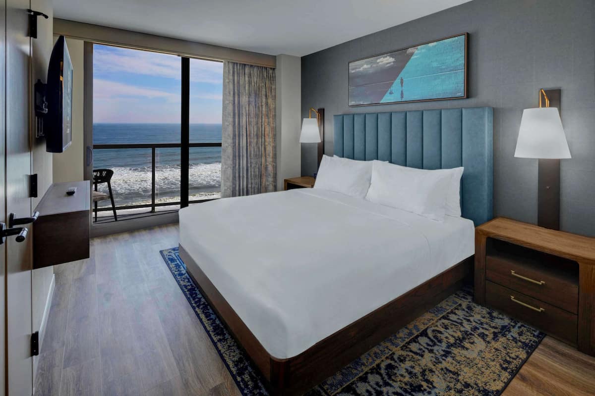 A hotel room with a large bed, a TV, a desk, and a view of the ocean.