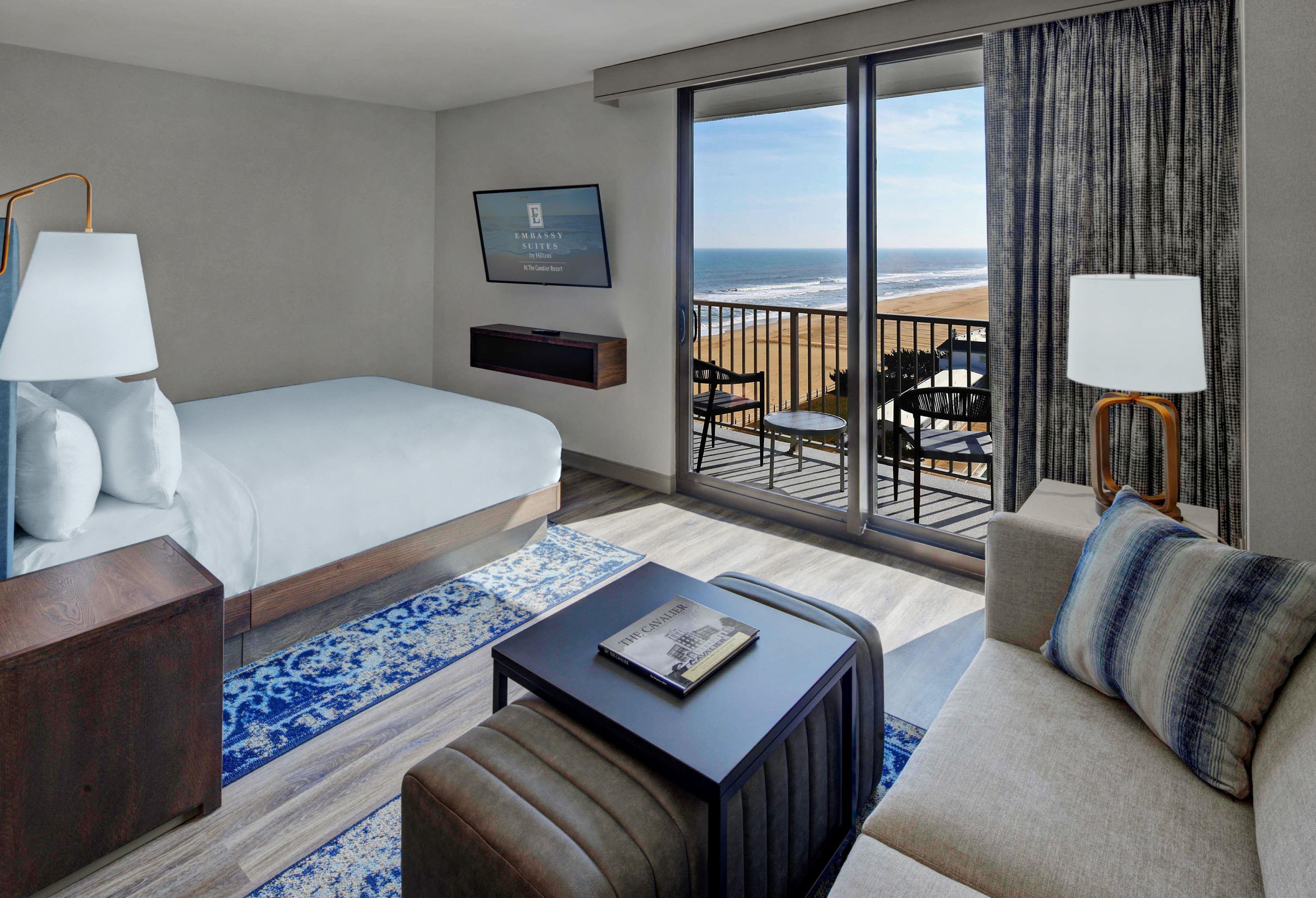 A hotel room with a bed, a sofa, a coffee table, a TV, and a balcony with a view of the beach.