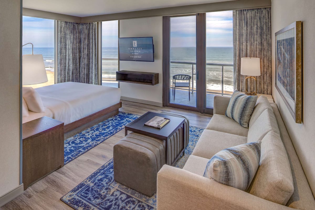 A hotel room with a bed, a sofa, a coffee table, a TV, and a balcony with a view of the ocean.