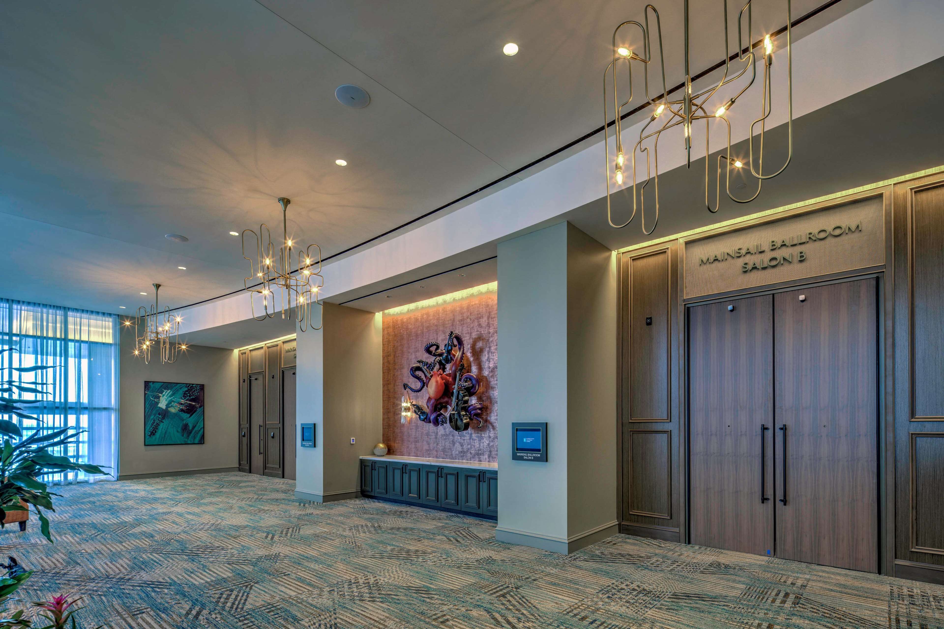 Un hall d’hôtel spacieux, doté d’un tapis à motifs, d’un comptoir de réception, d’une grande œuvre d’art murale et d’un panneau indiquant l’entrée du salon Mainsail Ballroom, salle B.