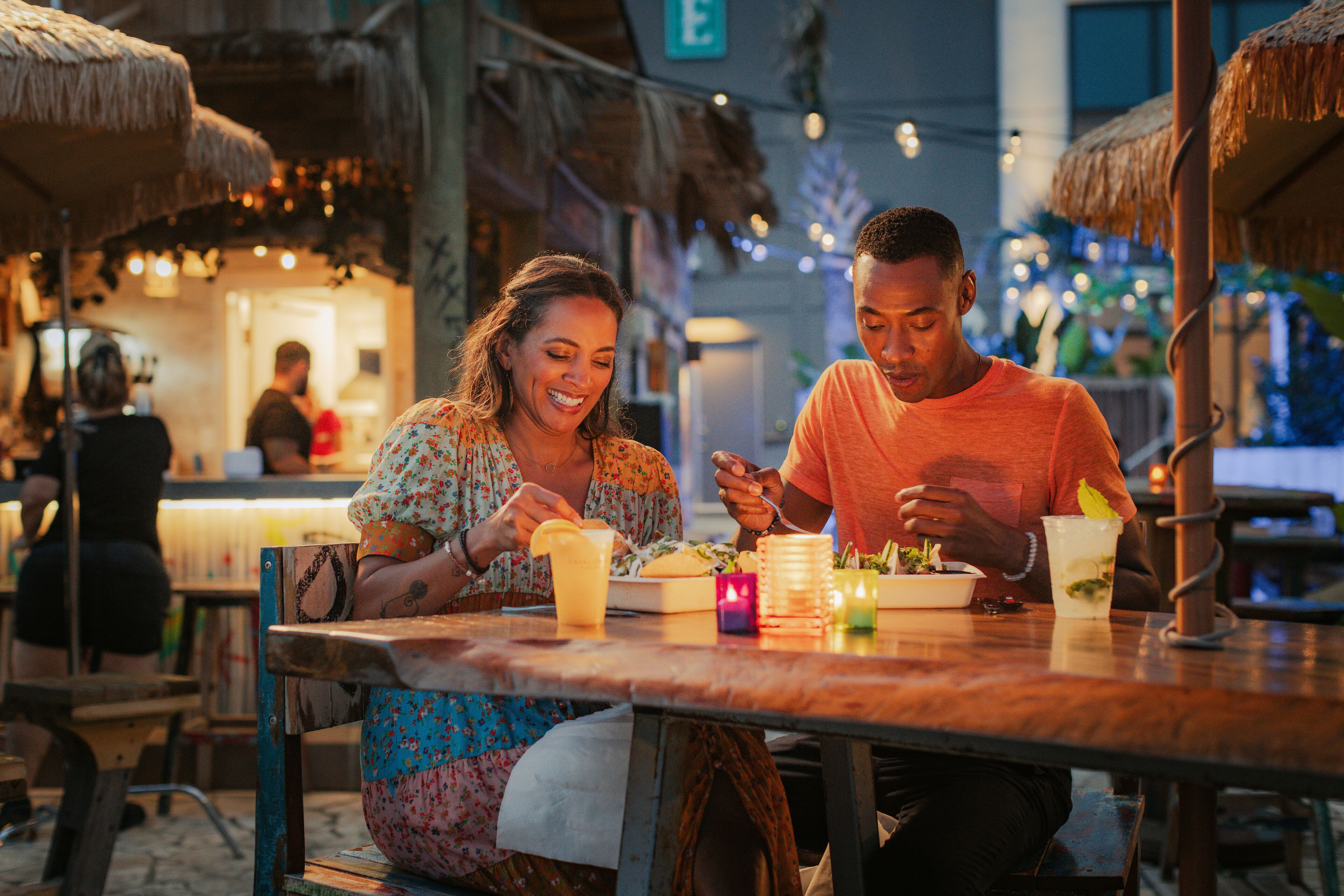 Una pareja cenando en un restaurante de temática tropical, con sombrillas de paja y luces colgantes.