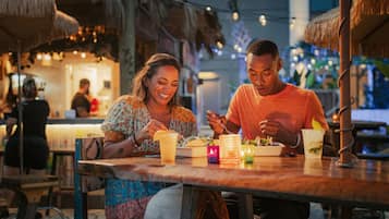 A couple dining at a restaurant with a tropical theme, featuring thatched umbrellas and string lights.