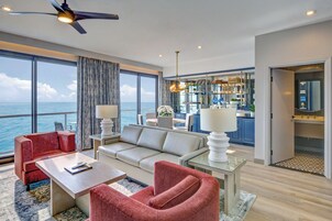 Una sala de estar moderna con sofá, sillones y una mesa de centro. Se ve el océano a través de amplias ventanas.