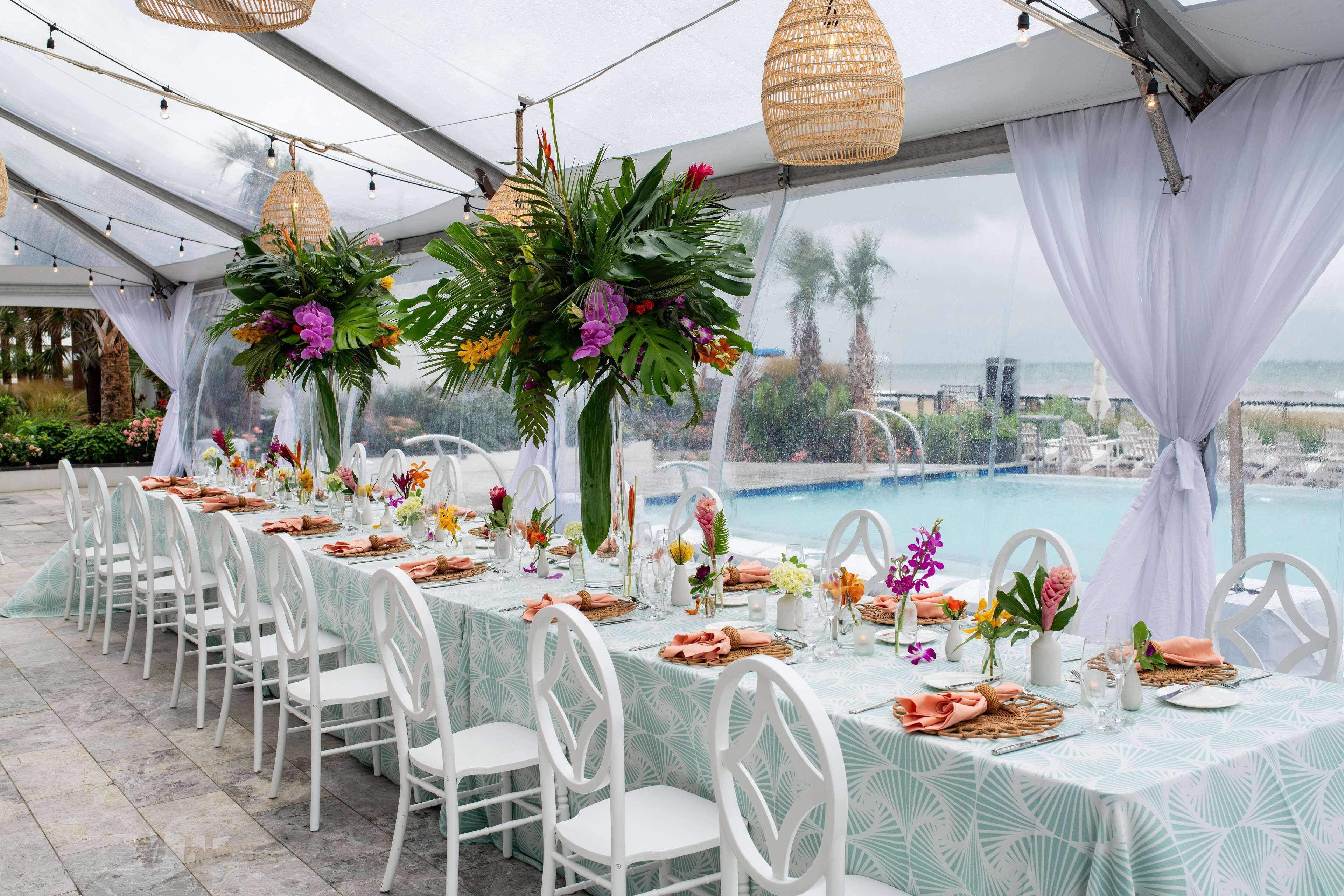 A tented outdoor dining area with tables set for a meal, surrounded by lush greenery and overlooking a pool.