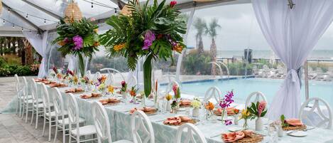 A tented outdoor dining area with tables set for a meal, surrounded by lush greenery and overlooking a pool.