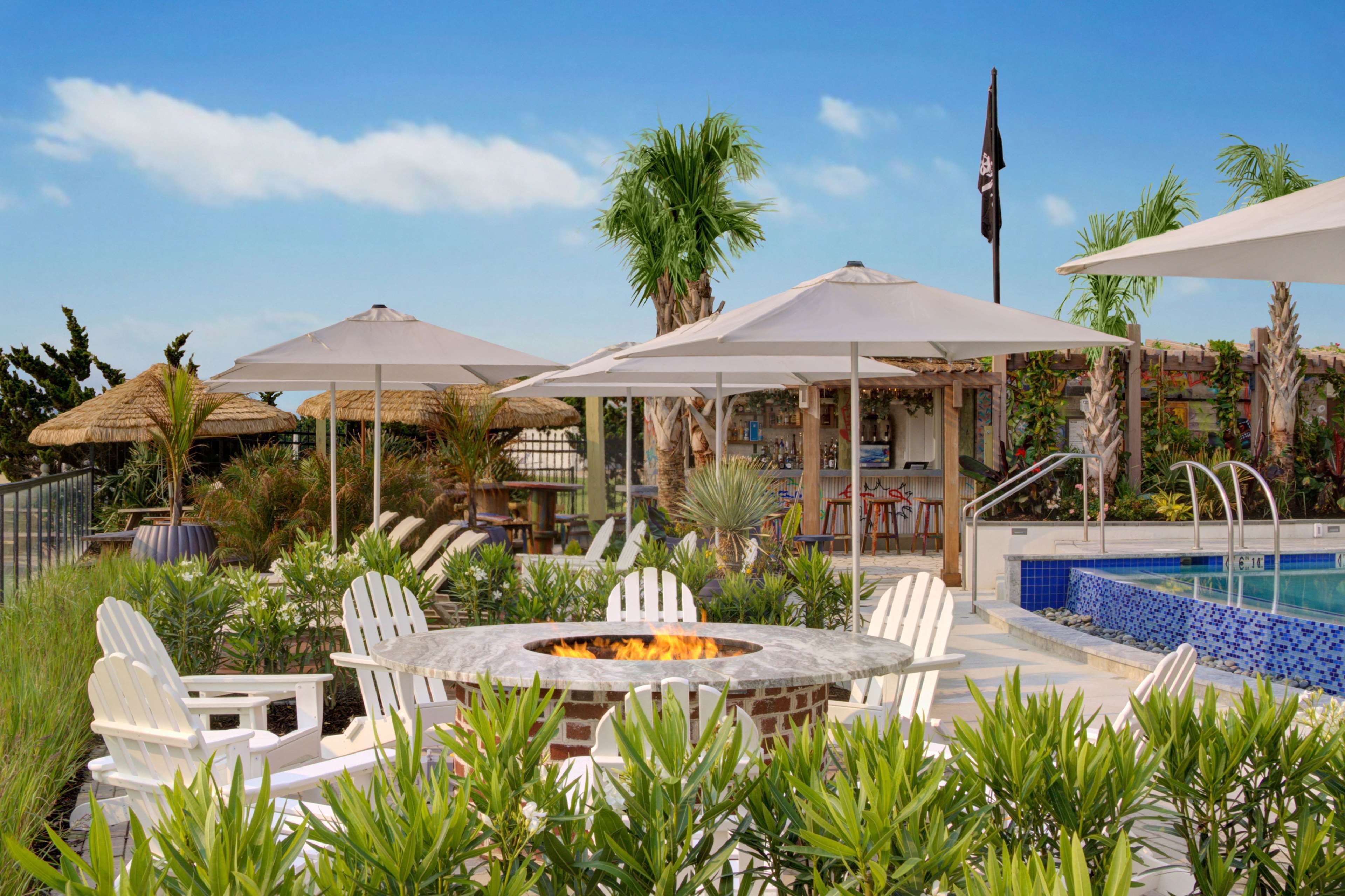 A poolside area with a fire pit, white Adirondack chairs, and umbrellas.