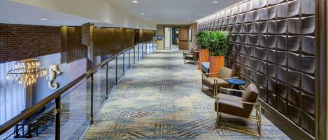 A hotel corridor with a patterned carpet, a glass railing, seating area with chairs and a table, and a wall with a textured design.