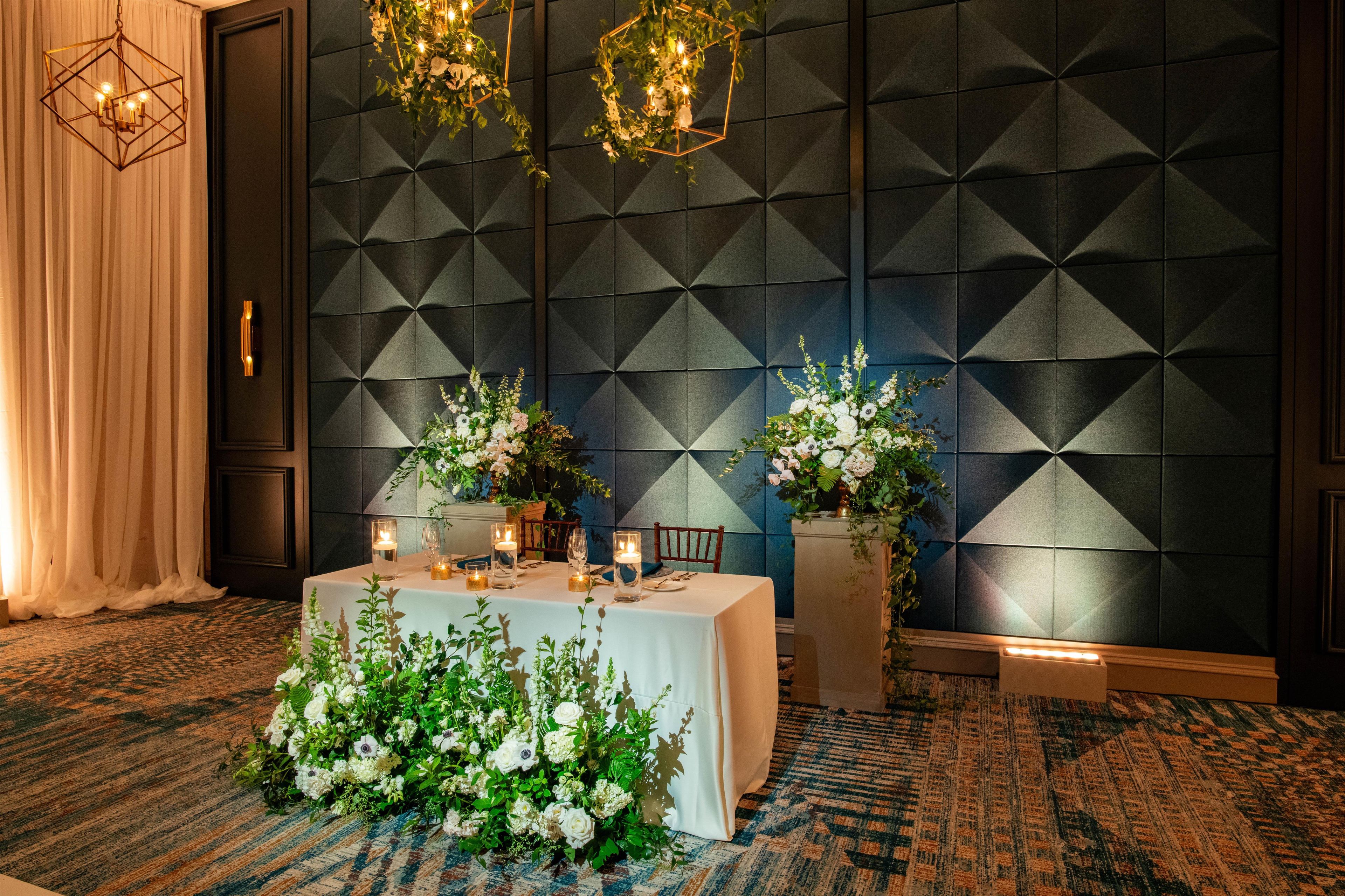 Une chambre d’hôtel avec un mur orné d’un motif géométrique, une table décorée de bougies et de fleurs, et un grand arrangement floral posé sur le sol.